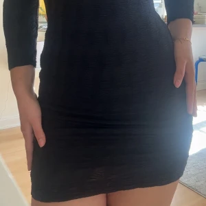 Svart tight kortklänning med v-ringning - Säljer en svart tight kortklänning med v-ringning och trekvartsärm. Klänningen har en diskret struktur i tyget och sitter figurnära. Perfekt för dig som gillar en enkel men stilren look.