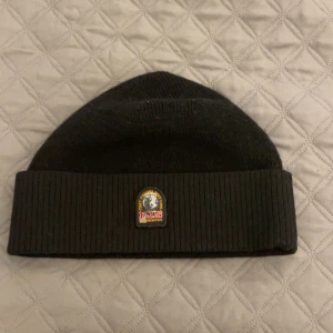 Svart stickad mössa från parajumper - Svart stickad mössa med bred uppvikt kant och ett broderat parajumper-märke framtill. Klassisk och enkel design som passar till det mesta. Perfekt för kyliga dagar.