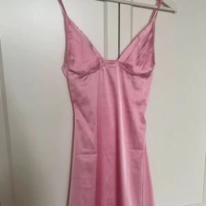 Rosa satinklänning med smala axelband - Säljer en söt rosa satinklänning med smala axelband och lätt v-ringning. Klänningen har en figurnära passform upptill och faller fint nedåt. Perfekt för sommarens tillfällen när du vill känna dig extra fin. Endast testad