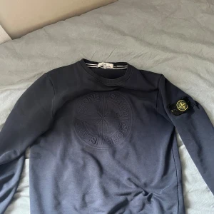 Mörkblå sweatshirt från Stone Island - Säljer en mörkblå sweatshirt från Stone Island med klassisk logga på ärmen och broderad logga på bröstet. Tröjan har rund hals och lång ärm, perfekt för en stilren look.