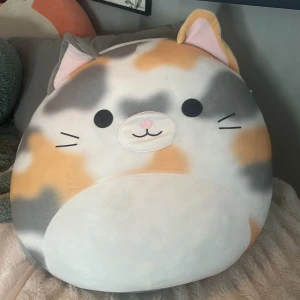 Stor katt Squishmallow - Supermjuk och rund kattgossedjur i form av en katt från Squishmallows. Katten har grå, vit och orange päls med söta öron, svans och broderat ansikte. Perfekt som gosedjur eller mysig inredningsdetalj.Orginal pris 800kr. Pris kan diskuteras!