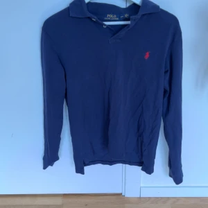 Marinblå långärmad pikétröja från Polo Ralph Lauren - Säljer en klassisk marinblå långärmad pikétröja från Polo Ralph Lauren med röd broderad logga på bröstet. Tröjan har krage och knappar vid halsen, perfekt för en stilren look. M i storlek men funkar som S