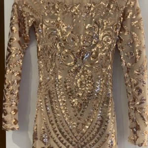 Glittrig kortklänning med paljetter - Säljer en superglittrig kortklänning med långa ärmar och snyggt mönster av guldiga paljetter. Klänningen har en tight passform och är perfekt för dig som vill sticka ut. Materialet är syntet och den har en nude/beige bas.