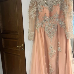 Långklänning i persikofärgad tyll med silverbroderier - Elegant långklänning i persikofärgad tyll med glittriga silverbroderier och pärlor. Klänningen har långärmade meshärmar och vacker öppen rygg. Perfekt för bal eller fest. Säljes då den inte längre kommer till användning.