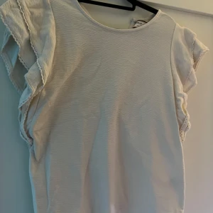 Vit volangtopp från Holly & Whyte - Säljer en söt vit topp från Holly & Whyte med volangdetaljer längs ärmarna. Toppen har rund halsringning och är tillverkad i ett lätt strukturerat material. Perfekt för en fräsch och stilren look.