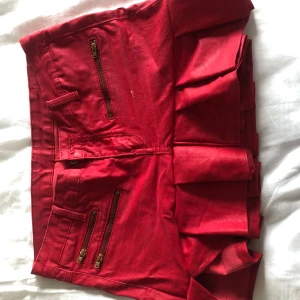 Röd minikjol med volang - Säljer en röd minikjol med volangkant och dragkedjedetaljer. Kjolen har två fickor bak och två fickor fram med guldiga dragkedjor. Perfekt för dig som vill sticka ut med en färgstark look. Med slits😍