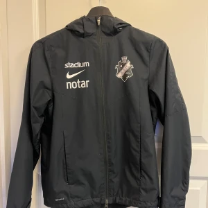 Svart AIK vindjacka från Nike - Svart vindjacka från Nike med AIK-logga på bröstet och tryck från Stadium, notar och SVEA. Jackan har huva, dragkedja framtill och två sidofickor. Perfekt för fotbollsträning eller som supporterjacka. Luvan är avtag bar. Strl är L i barnstorlek vilket motsvarar 147-158 cm lång, se sista bild. Hör av er vid frågor👍