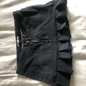 Mörkblå jeanskjol med volang från Lion Hundred Jeans - Supersnygg mörkblå jeanskjol med dragkedja framtill och volangdetalj längs nederkanten. Kjolen har en kort passform och är perfekt för dig som gillar en trendig och lekfull stil.