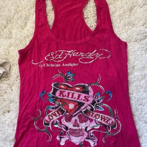 Rosa linnetopp från Ed Hardy - Säljer en starkt rosa linnetopp från Ed Hardy med tryck av dödskalle, hjärta och texten 'Love Kills Slowly' framtill. Toppen har brottarrygg och glittriga detaljer i trycket. Perfekt för dig som gillar statement-plagg!