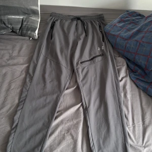 Grå byxor från Under Armour - Säljer ett par grå träningsbyxor från Under Armour med dragsko i midjan och dragkedjeficka på höger sida. Byxorna har en sportig look och är perfekta för träning eller chill. det står XL på dom men dom är L passar samt M