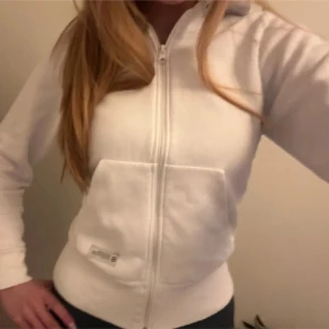 Vit hoodie från Stronger - Säljer en vit hoodie från Stronger med dragkedja och fickor framtill. Tröjan har huva och logga på bröstet samt ryggen. Perfekt för en avslappnad stil och enkel att matcha med det mesta.