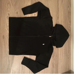 Svart hoodie Polo Ralph Lauren - Svart hoodie med dragkedja från Polo Ralph Lauren. Klassisk design med huva och vit broderad logga på bröstet. Mjuk bomullskvalitet och fickor framtill. Perfekt för en avslappnad stil.