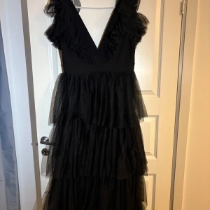 Balklänning - Säljer En svart långklänning med djup v-ringning och flera lager volanger i tyll. Klänningen har breda axelband med volangdetaljer och en luftig. Aldrig använd men prislappen finns tyvärr inte kvar. 