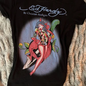 Svart Ed Hardy t-shirt med färgglatt tryck - Bud!!!   Högsta bud just nu är 500😊                          Svart t-shirt från Ed Hardy med stort färgglatt tryck av en tecknad pinup-tjej och grön fjäder på framsidan. T-shirten har rund halsringning och korta ärmar. Ed Hardy-logga i blått både fram och bak. Äkta och vintage från min mamma som jag fått ärva . Storlek XL men är absolut inte så stor och passar mig som är S. 
