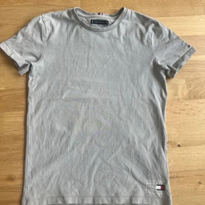 Ljusgrå t-shirt från Tommy Hilfiger - Snygg ljusgrå t-shirt från Tommy Hilfiger med klassisk rund hals och diskret logga samt tryck nertill på framsidan. T-shirten är enkel och stilren, perfekt till jeans eller shorts. Storleken är dock oklar men skulle tro att det är ca 165-168cm.