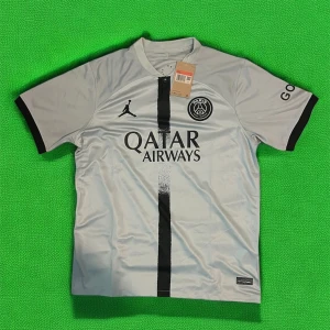 PSG fotbollströja med Qatar Airways sponsor - Snygg vit PSG fotbollströja med svart vertikal rand, Jordan-logga och Paris Saint-Germain klubbmärke på bröstet. Svarta detaljer på ärmslut och tryck på ärmen. Perfekt för fotbollsälskare!