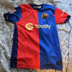 FC Barcelona hemmatröja Lamine Yamal 27 - Säljer en officiell FC Barcelona fotbollströja med Lamine Yamal och nummer 27 på ryggen. Tröjan är röd och blå med klubbmärke, Nike-logga och Spotify som sponsor framtill. Kortärmad modell i lätt och ventilerande material.