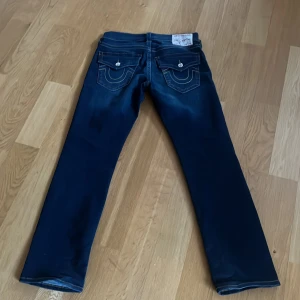 Mörkblå jeans från True Religion - Säljer ett par mörkblå jeans från True Religion med klassiska kontrastsömmar och snygga fickdetaljer bak. Jeansen har raka ben och låg midja, samt dekorativa knappar på bakfickorna. Perfekta för dig som gillar streetwear och vill sticka ut. Storlek 30