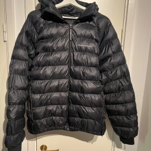 Svart dunjacka från Peak Performance - Säljer en svart dunjacka från Peak Performance med huva och dragkedja framtill. Jackan har en quiltad design och logga på bröstet samt ärmen. Perfekt inför vår sommar och höst dagar och enkel att matcha med olika outfits. 2 små sydda defekter se bild. Justerbar luva samt dragkedja vid fickorna. Mått - Längd 70cm, Bredd (axlar) 67cm, Bredd (Midja) 58cm