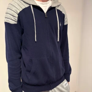 Marinblå hoodie med grå huva från Ralph Lauren - Säljer en marinblå hoodie från Ralph Lauren med grå huva och axelparti. Tröjan har dragkedja framtill, två fickor och långa ärmar. Perfekt för en avslappnad stil och enkel att matcha med andra plagg. Material merino ull och kashmir
