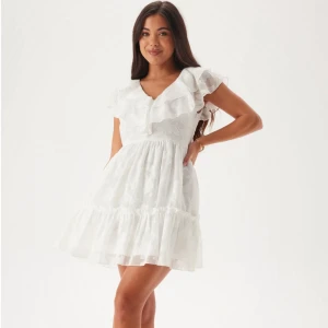 Vit volangklänning med v-ringning - Söt vit kortklänning med volangdetaljer och v-ringning. Klänningen har korta ärmar och en luftig passform, perfekt för varma dagar. Tillverkad i ett lätt material som ger en romantisk känsla. Perfekt till student eller konfirmation! Helt oanvänd med lappen kvar!