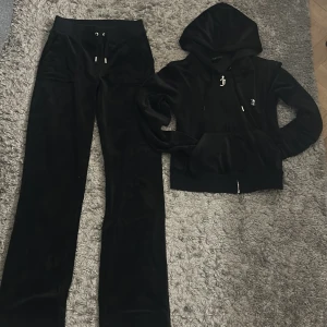 Svart velour hoodie från Juicy Couture - Säljer en svart velour hoodie från Juicy Couture med dragkedja och juicy print i glitter på ryggen. Säljer även ett par svarta velour byxor från juicy Couture med fickor där bak. Det finns en liten defekt på tröjan där en metallbit på snöret har gått av.
