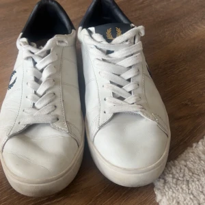 Vita sneakers från Fred Perry - Snygga vita sneakers från Fred Perry med klassisk design. Skorna har en svart logotyp på sidan och vita snören. Perfekta för en stilren look.