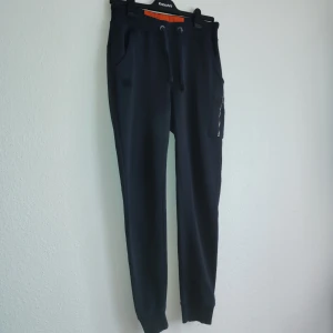 Svarta mjukisbyxor från Alpha Industries - Snygga svarta mjukisbyxor från Alpha Industries med dragkedja och snörning i midjan. Byxorna har en praktisk ficka med dragkedja på sidan och är perfekta för en avslappnad stil. De är tillverkade i ett mjukt och bekvämt material.