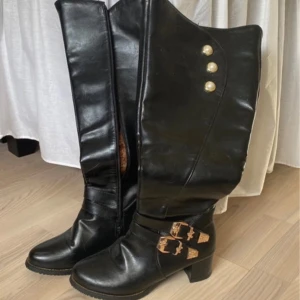 Svarta stövlar med leopardmönstrad insida - Snygga svarta stövlar i skinn med leopardmönstrad insida. De har dekorativa guldfärgade spännen och knappar samt en dragkedja på sidan. Perfekta för en trendig look. Och helt nya aldrig använd 