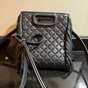 Maje- Svart axelväska med fransar  - Snygg svart axelväska från Maje med quiltad design och fransar på sidorna. Väskan har en praktisk axelrem och små nitar som ger en extra touch. Perfekt för att ge din outfit en cool och trendig look.