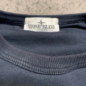 Mörkblå tröja från Stone Island Junior - Säljer en snygg mörkblå tröja från Stone Island Junior. Tröjan har långa ärmar och en klassisk rund halsringning. Den har en ikonisk logga på ärmen som ger en cool touch. Perfekt för en stilren look. Stl 150-160