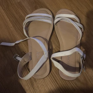 Vita sandaler med remmar - Snygga vita sandaler med flera remmar och spänne runt ankeln. Perfekta för varma sommardagar och ger en stilren look. Bekväma att bära och lätta att matcha med olika outfits.