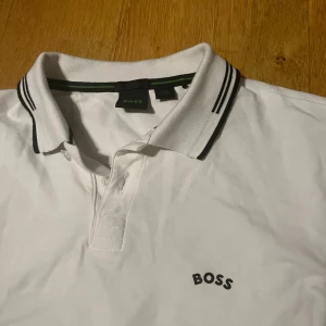 Vit pikétröja från Boss - Säljer en stilren vit pikétröja från Boss med klassisk krage och knappar. Tröjan har svarta detaljer på kragen och ärmsluten samt en diskret Boss-logga på bröstet. Perfekt för en avslappnad men ändå snygg look. Storlek M. Köpt för 899 men säljer för 599