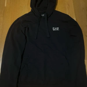 Svart hoodie från Emporio Armani - Säljer en stilren svart hoodie från Emporio Armani med EA7-logga på bröstet. Tröjan har en klassisk känguruficka och justerbar huva med snören. Perfekt för en avslappnad look. Storlek M men sitter lite tight. Knappast andvänd. Köpt för 999 men säljer för 599