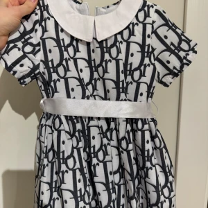 Vit och svart klänning med logotypmönster - Söt klänning med korta ärmar och ett iögonfallande svart logotypmönster på vit bakgrund. Den har en vit krage och ett brett satinband i midjan som knyts i en rosett bak. Perfekt för små fashionistas! Storlek 80-86