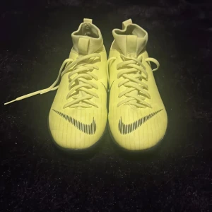 Gula fotbollsskor från Nike - Snygga gula fotbollsskor från Nike med snörning och hög ankel. Skorna har en modern design med svarta detaljer och en slitstark sula för bra grepp på planen. Perfekta för den som vill sticka ut på fotbollsplanen.