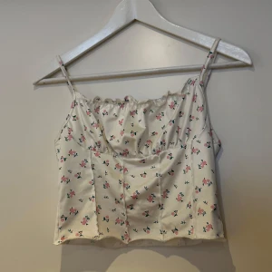 Blommig vit linnetopp - Söt vit linnetopp med små blommor i rosa och blått. Toppen har smala axelband och en lätt rynkad detalj vid halsringningen. Perfekt för varma sommardagar! Linnet är från shein