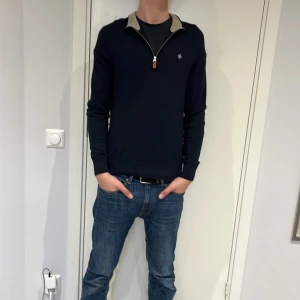 Svart tröja i merinoull från Morris - Snygg svart tröja från Morris i 100% merinoull. Tröjan har en dragkedja vid halsen och en kontrasterande krage i beige. Perfekt för en stilren look.