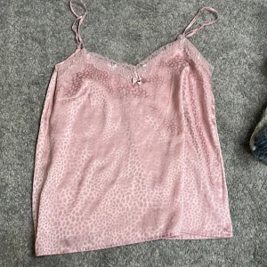 Rosa satinlinne med spetsdetaljer - Säljer ett sött rosa satinlinne med tunna axelband och spetsdetaljer vid halsringningen. Linnet har ett diskret leopardmönster och en liten rosett med ett hjärthänge framtill. Perfekt för en romantisk look!