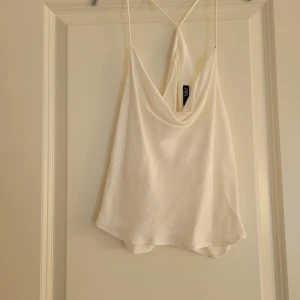 Vit topp från Zara - Säljer en stilren vit topp från Zara med tunna axelband och draperad framsida. Perfekt för varma sommardagar eller som ett lager under en kavaj. Toppen har en luftig och bekväm passform.