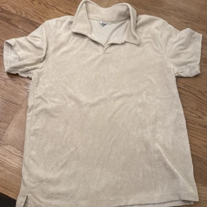 Beige frotté t-shirt strlk 170 - Säljer en beige t-shirt i frotté med korta ärmar och krage. Perfekt för en avslappnad stil. Hör av dig vid intresse!