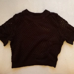 Svart stickad topp - Säljer en svart stickad topp med korta ärmar och ett luftigt hålmönster. Perfekt för lager-på-lager eller som en stilren detalj i garderoben. Toppen är croppad och har en rund halsringning.