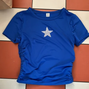 Blå topp med stjärndetalj från Shein - Säljer en blå topp från Shein med en glittrig stjärna på bröstet. Toppen har korta ärmar och rynkade sidor för en snygg look. Perfekt för att lägga till lite bling i vardagen!