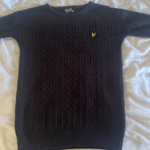 Mörkblå  stickad tröja från Lyle & Scott - Snygg mörkblå stickad tröja från Lyle & Scott med kabelstickat mönster och deras ikoniska gula logga på bröstet. Perfekt för kyligare dagar med sin klassiska design och bekväma passform.