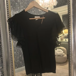Svart topp från Holly & Whyte - Snygg svart topp från Holly & Whyte med volangdetaljer på ärmarna. 