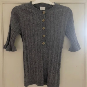 Grå ribbad kofta från Mamalicious - Säljer en stilren grå ribbad kofta från Mamalicious. Koftan har trekvartsärmar med volangdetaljer och knappar framtill. Perfekt för lager-på-lager under kyligare dagar.