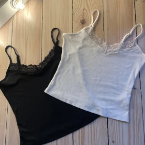 Svart och vit ribbad linnetopp med spetsdetaljer - Två snygga ribbade linnen i svart och vitt med spetsdetaljer vid halsringningen. Perfekta för att lägga till en touch av elegans till din outfit. De har smala axelband och en mjuk, bekväm passform. Aldrig använda.