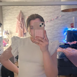 Vit ribbad topp med volangärmar - Säljer en söt vit ribbad topp med volangdetaljer på ärmarna. Perfekt för en stilren look. Toppen har en rund halsringning och är i ett mjukt material. Toppen har en svag parfymfläck på framsidan som jag ej lyckats få bort i tvätten (se sista bilden).