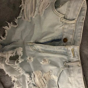 Ljusa jeansshorts med slitningar - Säljer ett par ljusa jeansshorts med coola slitningar och fransiga kanter. Perfekta för en avslappnad stil. De har en knapp och dragkedja framtill samt fickor både fram och bak.