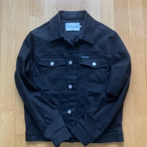 Svart jeansjacka från Calvin Klein - Snygg svart jeansjacka från Calvin Klein med stora vita bokstäver på ryggen. Jackan har en klassisk design med knappar framtill och två bröstfickor med lock. Perfekt för en stilren look!